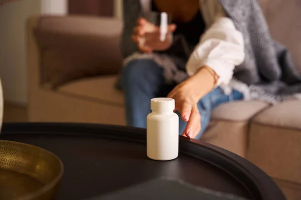 Woman sits with a glass of water on the sofa, she reaches to the table for a bottle of pills