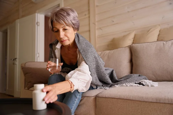 Ill pensioner sits with glass of water and jar of pills on sofa, she is wrapped in a warm shawl