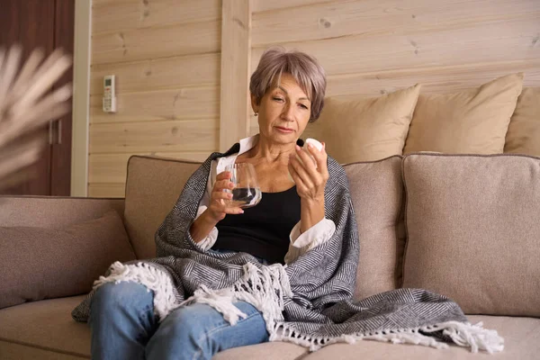 Sick woman sits with a glass of water and a jar of pills on the sofa, she is wrapped in a warm shawl