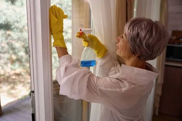 Woman with lilac hair washes a window with a yellow rag, a woman works in protective household gloves