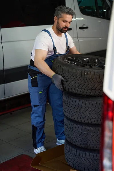 Focused auto repair worker lifting heavy car tyre while standing near van in garage