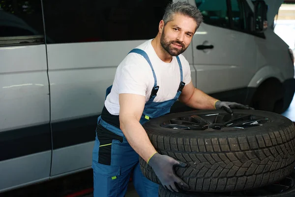 Auto mechanic lifting heavy rubber car tire while standing near motor vehicle at service station