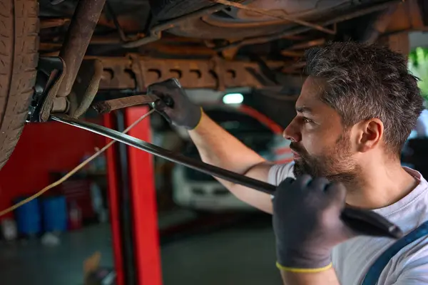 Serious auto mechanic using prybar and lamp for inspecting car suspension system