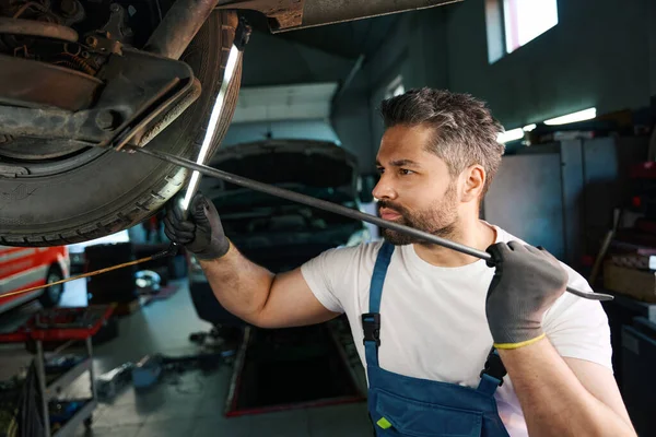 Serious concentrated auto mechanic checking car suspension parts using lamp and prybar