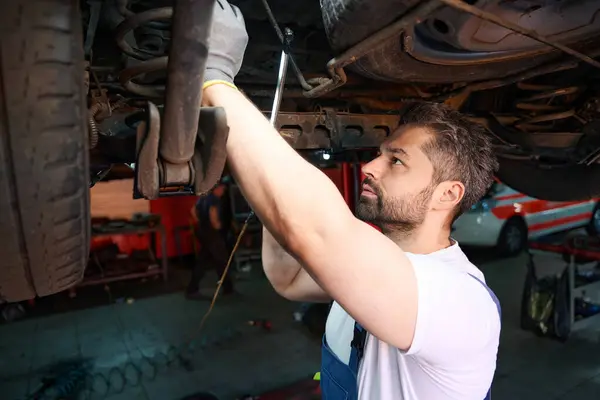 Serious focused automotive service technician checking car suspension parts using inspection lamp