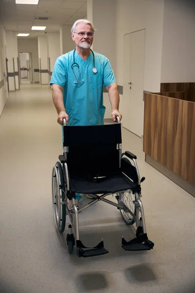 Serious medical worker leaning on wheelchair push handles in inpatient facility corridor