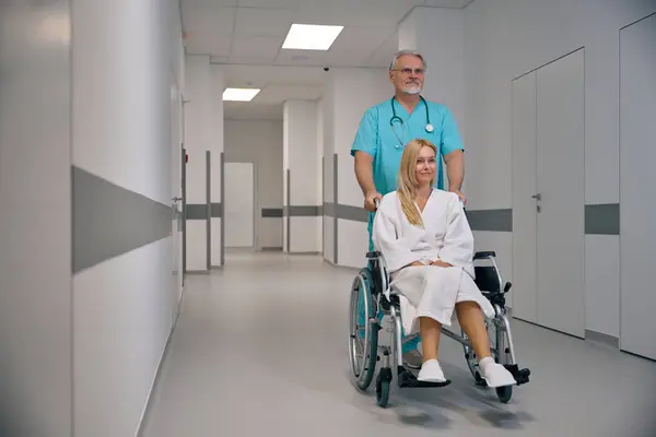 Mature healthcare worker pushing wheelchair with disabled female patient along hospital corridor