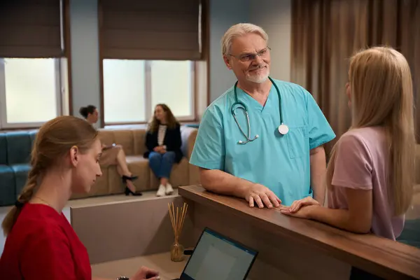Male doctor leaning on reception desk talking to out-patient in presence of busy receptionist