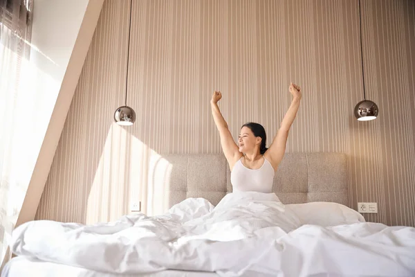 Sleepy woman seated in comfortable bed in suite stretching herself while looking into distance