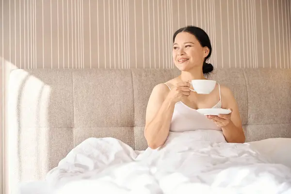 Smiling female with cup of coffee and saucer in hands seated in bed in suite and looking away