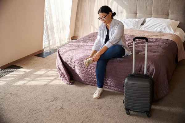Exhausted female traveler seated on bed in suite pulling off her sneakers