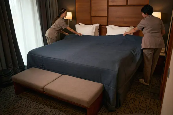 Two chambermaids dressed in uniforms making bed with bedspread in hotel room