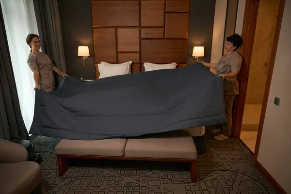 Cheerful maid in uniform and her colleague making bed with bedspread in hotel room