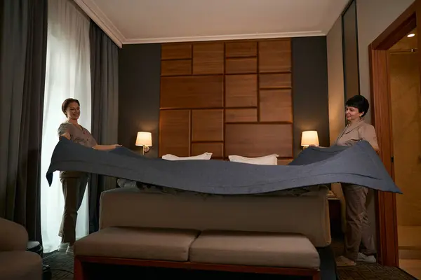 Smiling uniformed maid service worker making bed with bedcover assisted by her colleague
