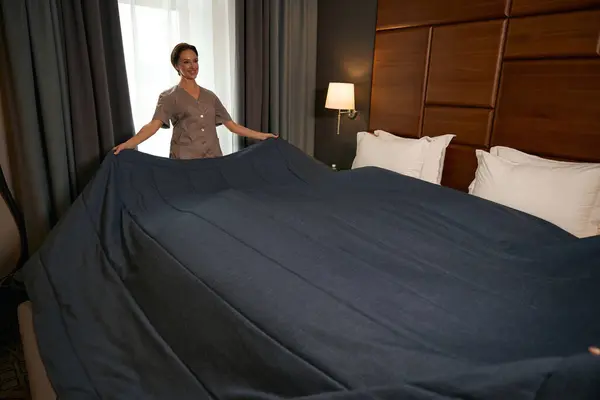 Smiling uniformed maid service worker making bed with bedspread in hotel room