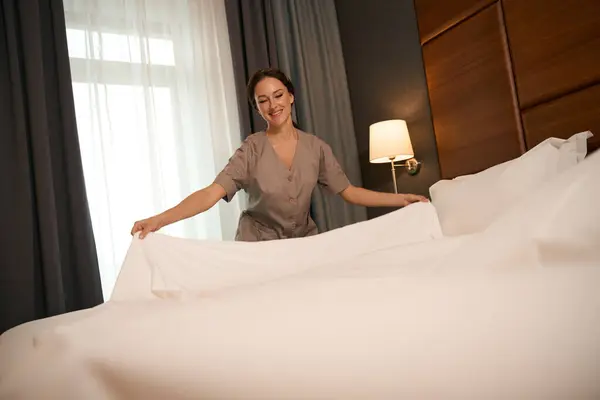 Contented chambermaid dressed in uniform putting bedsheets on bed in hotel room