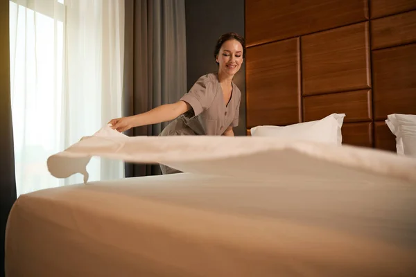 穿着制服的快乐的年轻女服务员在套间里用干净的羽绒做床 — 图库照片