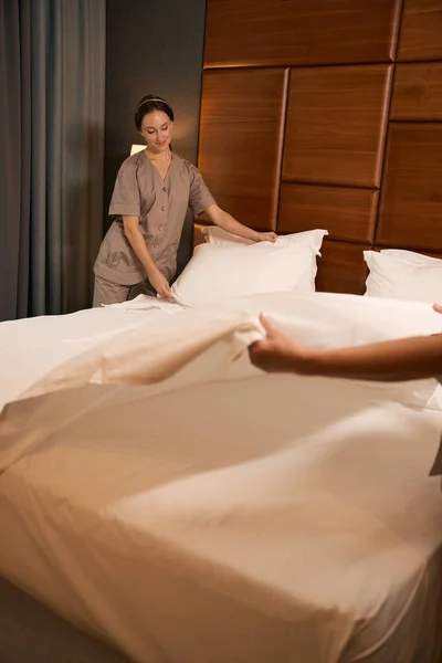 Pleased maid in uniform making bed in hotel room assisted by colleague
