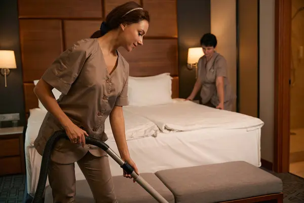 Smiling chambermaid in uniform vacuum-cleaning carpet in hotel room while her colleague making bed