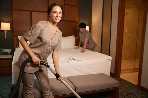 Joyous maid service worker vacuum-cleaning carpet in suite while her coworker making bed