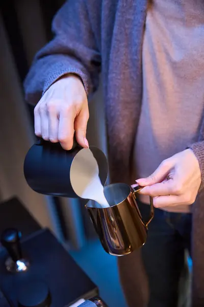 Unrecognizable woman barista pouring milk from pitcher preparing coffee in coffee shop