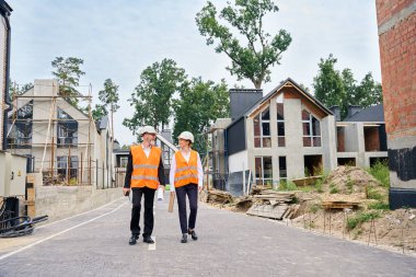 Full-size portrait of foreman with walkie-talkie and drawings talking to female colleague walking along street with half-constructed houses clipart