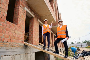 İnşaat müdürü ahşap merdivende duruyor. Müteahhitlere bir şey gösterirken bitmemiş evin dışında.