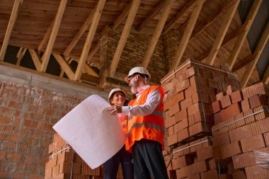 Foreman, tamamlanmamış bir evin çatısında dururken memnun kadın inşaat müdürünün planlarını gösteriyor.