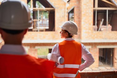 Kolunun altında katlanmış mimari çizimleri olan bir bina yöneticisi ustabaşının önünde tamamlanmamış tuğla eve bakıyor.