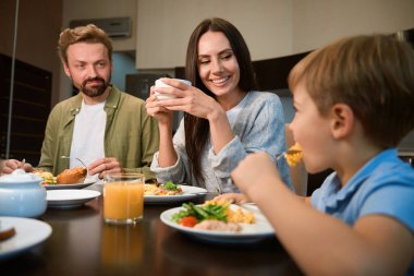 Otel odasında ailesiyle kahvaltı ederken sabah kahvesini içen gülümseyen kadın.