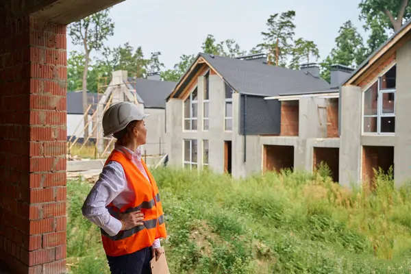 Competent female construction supervisor in safety vest and hard hat inspecting unfinished house exterior