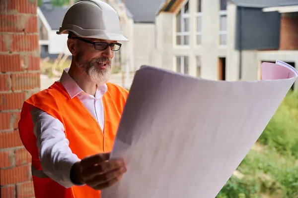 Waist-up portrait of contented foreman looking at blueprints in his hands while standing among half-built houses