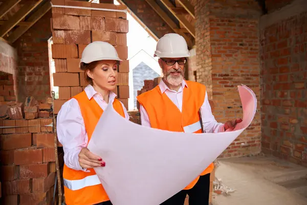 Focused building supervisor and contractor reading blueprints while standing in unfinished house
