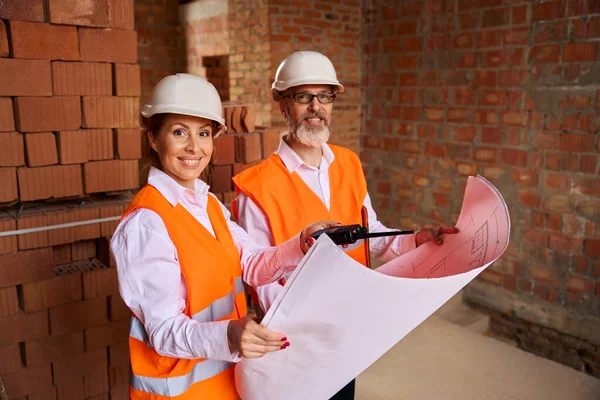 Smiling building inspector and foreman with blueprints in hands while standing among brick walls of unfinished house