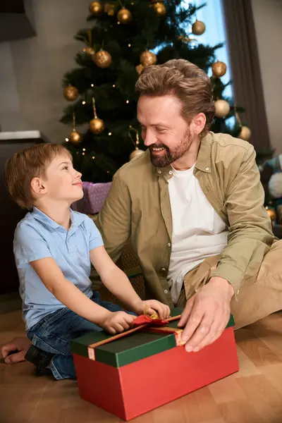 Father and son opening Christmas present box celebrating New Year in hotel sitting near Xmas tree