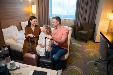 Anne, baba ve kızı, bavulları yataklarında oturmuş birbirlerine bakıyorlar. Dinlenme, tatil ve seyahat kavramı. Aile ilişkisi ve birlikte zaman geçirme fikri