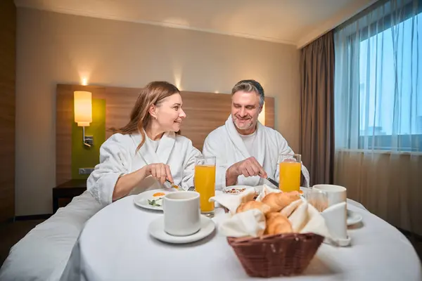 笑顔の大人の慎重なカップルは ホテルの部屋のテーブルでベッドで朝食をとるバスローブを着ています 旅行の概念 — ストック写真