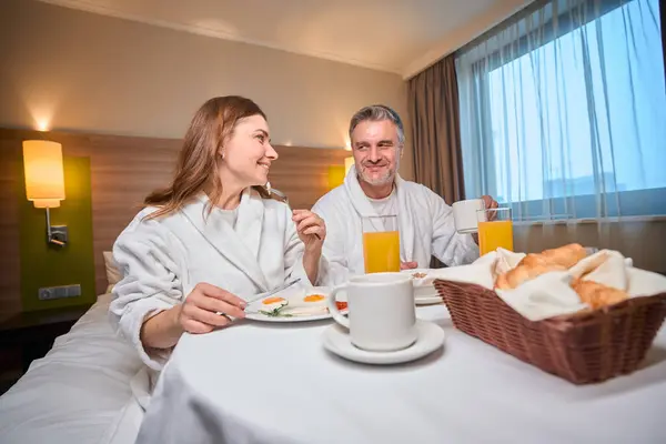笑顔の大人の慎重なカップルは朝食を持って ホテルの部屋のテーブルのベッドでお互いを見ています 旅行の概念 — ストック写真