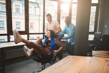 Erkek, çok ırklı iş arkadaşları, ofis etrafında tekerlekli sandalyede örülmüş saçları olan, başarılı projenin bitişini kutlayan, iş arasında dinlenen, yuvarlanan kadın meslektaşlarıyla eğleniyor.