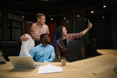 İş dünyasının dost canlısı iş arkadaşları akıllı telefonda poz veriyor ve selfie çekiyorlar, zaferi ve başarılı proje tamamlanışını kutluyorlar, çok ırklı takım rahat bir çalışma alanında çalışıyor.