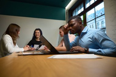 Diğer meslektaşlar birbirleriyle konuşurken, dikkatlerini dizüstü bilgisayarında yaptığı projeye vermeye çalışan Afrikalı Amerikalı bir adam.