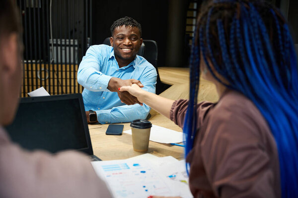 Smiling African American boss greeting and shaking hand to braided woman crisis manager who engaged in company development and money and power increasing, team working in comfortable office