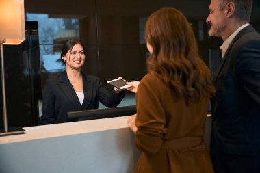 Gülümseyen Avrupalı kadın resepsiyonist otel lobisinde kayıt sırasında beyaz adamın yanında yetişkin bir kadından pasaport alıyor. Dinlenme, tatil ve seyahat kavramı
