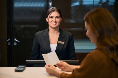 Yetişkin bir kadın otel lobisindeki resepsiyon sırasında gülümseyen Avrupalı kadın resepsiyonistin yanında pasaportunu izliyor. Dinlenme, tatil ve seyahat kavramı