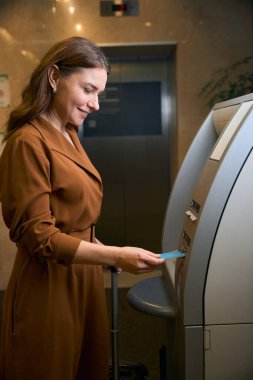 Elinde bavuluyla gülümseyen beyaz kadın otel lobisindeki ATM 'ye kredi kartı yerleştiriyor. Dinlenme, tatil ve seyahat kavramı