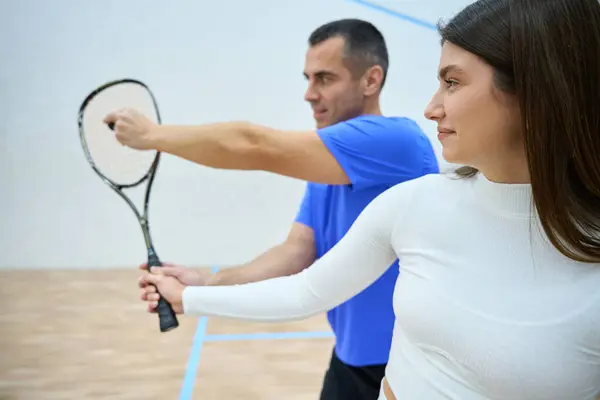 Sportive man instructor teaching woman squash emphasizing hitting skills on indoor court