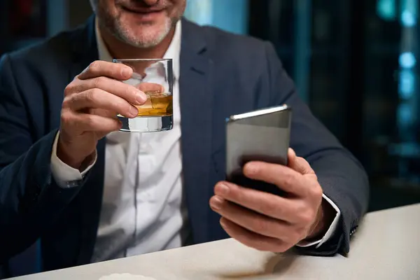 Obscure face of adult businessman using smartphone while drinking cognac from glass cup at reception desk in hotel lobby. Concept of rest, vacation and travelling