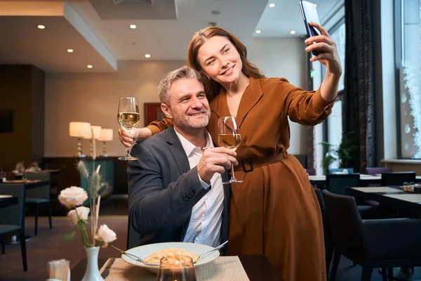 Adult happy caucasian couple with wine taking selfie on mobile phone in hotel restaurant. Concept of romantic date and event