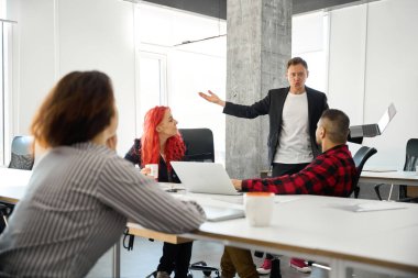 Eksantrik reklam yöneticisi laptopuyla sunumunu tam ofiste yapıyor, meslektaşları dikkatle dinliyor ve düzeltmeler öneriyor, beyin fırtınası