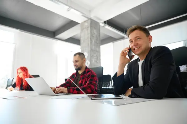 Confident man teamlead talking phone, arranging meeting with influential people who can help with business while his employees working on project on laptop in coworking space